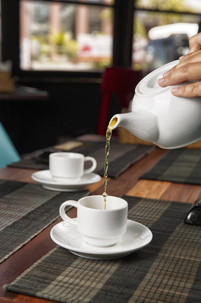 Giet thee uit een theepot in een kopje op een houten tafel, selectieve focus - Foto, afbeelding