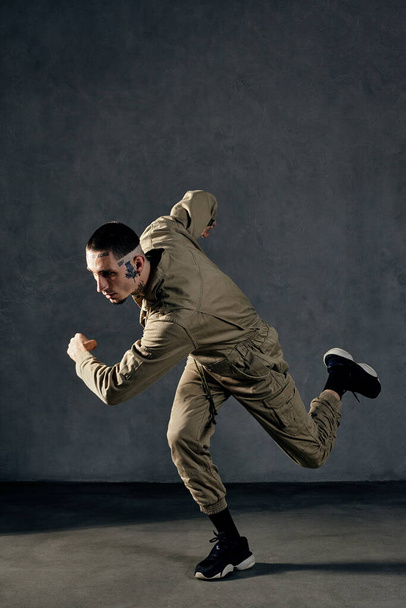Young stately fellow with tattooed body and face, earrings, beard. Dressed in khaki jumpsuit and black sneakers. He dancing against gray studio background. Dancehall, hip-hop. Full length, copy space - 写真・画像