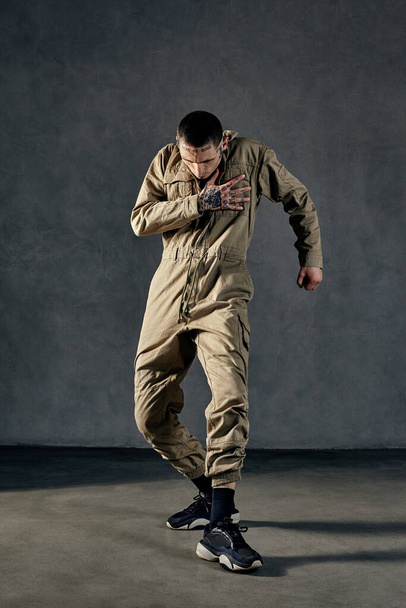 Young strong performer with tattooed body and face, earrings, beard. Dressed in khaki overalls and black sneakers. He is dancing against gray background. Dancehall, hip-hop. Full length, copy space - Photo, Image