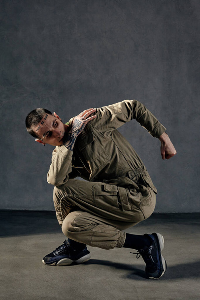 Extraordinary man with tattooed body and face, earrings, beard. Dressed in khaki overalls and black sneakers. He is dancing against gray studio background. Dancehall, hip-hop. Full length, copy space - Photo, Image