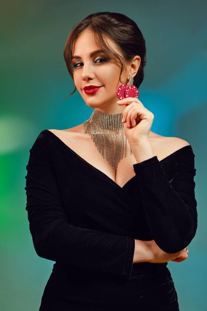 Brunette female with bare shoulders and bright make-up. In black dress, brilliant necklace and earrings. She is smiling, showing two red chips, posing on colorful background. Poker, casino. Close-up - Fotografie, Obrázek