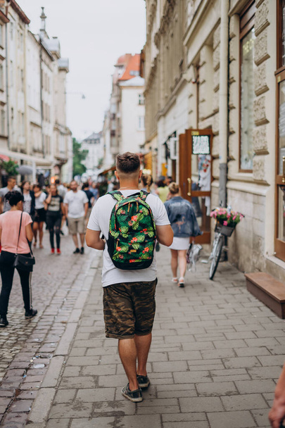 Muž s batohem na turné po městě v létě. Mladý muž chodí po městě za slunečného dne. Sportovní chlap v kraťasech a s barevným batohem - Fotografie, Obrázek