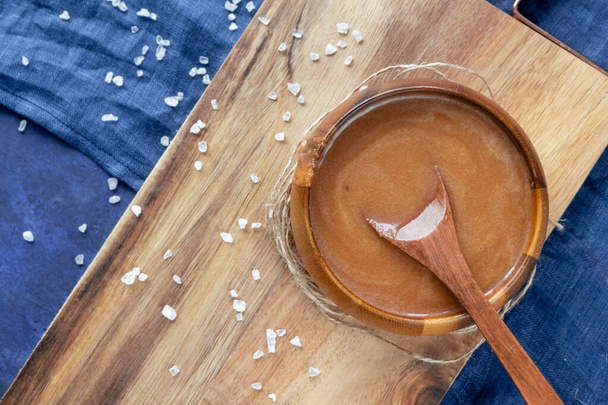  Homemade salted caramel sauce in a wooden bowl on a wooden board with a spoon, a sea salt and a blue textile background. View from above. - Foto, immagini