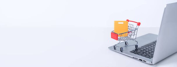 Online shopping. Mini shop cart trolley with colorful paper bags over a laptop computer on white table background, buying at home concept, close up - Fotoğraf, Görsel