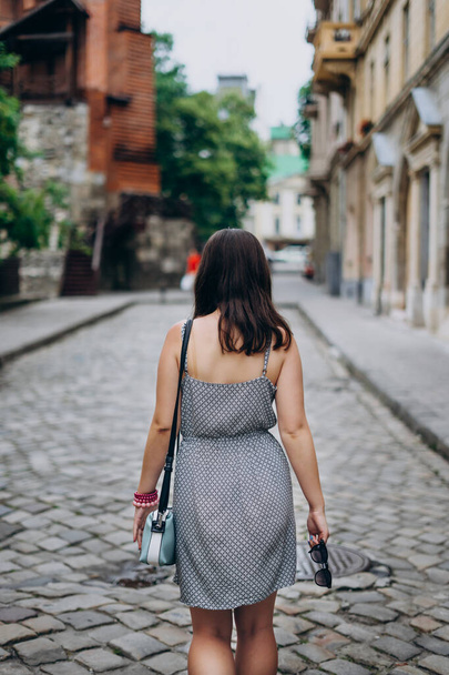 Young woman on a tour looks at beautiful homes in the city center. Girl in sundress with blue handbag and sunglasses. Girl walks the city in a dress. - Photo, Image