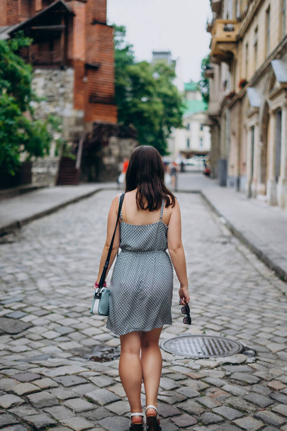 Mujer joven en un recorrido mira hermosas casas en el centro de la ciudad. Chica en vestido de sol con bolso azul y gafas de sol. Chica pasea por la ciudad en un vestido
. - Foto, imagen