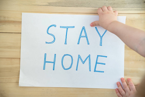 Kinderhand houdt papier met woorden STAY HOME op houten achtergrond. Quarantaine Covid-19. - Foto, afbeelding