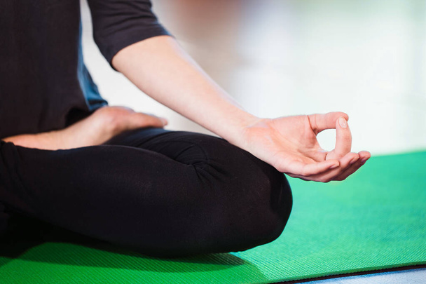Девушка с йоги медитирует в помещении и делает дзен-символ своей рукой. Крупный план женского тела в позе йоги на зеленом коврике
 - Фото, изображение
