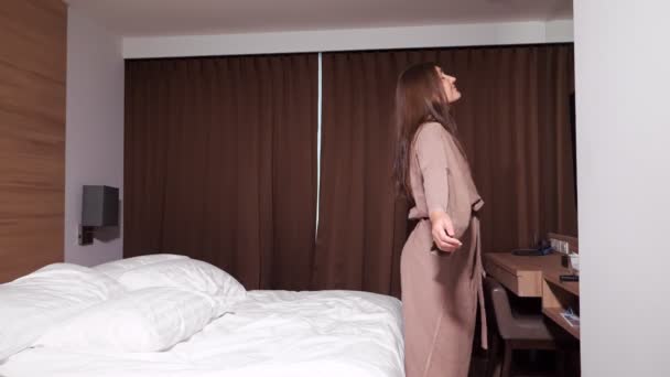 Nainen kylpytakissa hyppää sängylle hotellin ikkunaa vasten - Materiaali, video