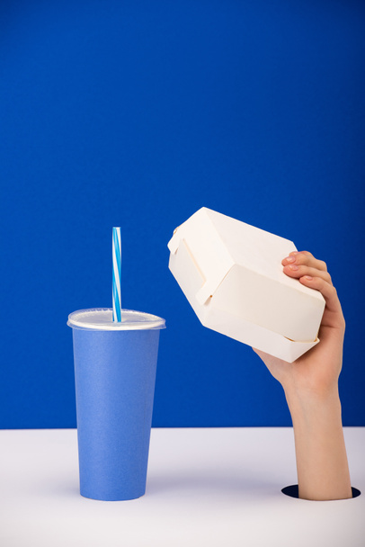 обрезанный вид женщины, держащей коробку рядом с бумажной чашкой с безалкогольным напитком изолированы на синий
 - Фото, изображение