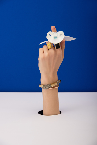cropped view of woman with bracelet and golden rings holding silicone pacifier isolated on blue - Φωτογραφία, εικόνα