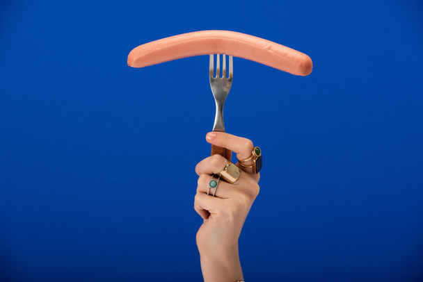bijgesneden weergave van vrouw met ringen op vingers vork met worst op blauw  - Foto, afbeelding