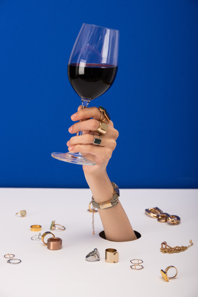abgeschnittene Ansicht einer Frau mit Armband an der Hand, die ein Glas mit Rotwein in der Nähe von Ringen hält, die auf blau isoliert sind - Foto, Bild