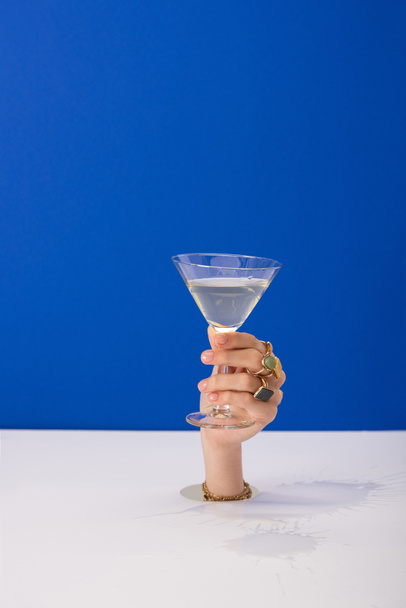 przycięty widok kobiety z bransoletką i pierścieniami trzymającymi szklankę koktajlu martini odizolowanego na niebiesko - Zdjęcie, obraz