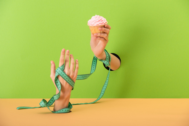 cropped view of woman with measuring tape on hands holding tasty cupcake on green and orange  - Foto, afbeelding