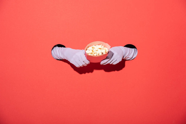 bijgesneden weergave van de vrouw in handschoenen houden emmer met lekkere popcorn op rood - Foto, afbeelding