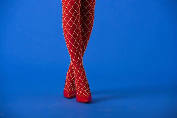 cropped άποψη της γυναίκας σε κόκκινο καλσόν fishnet και τακούνια στέκεται στο μπλε  - Φωτογραφία, εικόνα