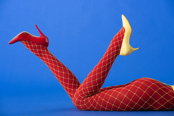 cropped view of model in bright fishnet tights, yellow and red heels lying on blue - Foto, Imagem