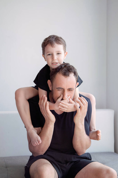 dad plays with his son in a bright room on a gray sofa near a large window, father's love - Photo, Image