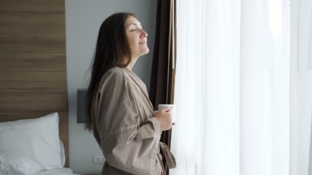 dívka v županu nápoje káva v blízkosti okna v hotelovém pokoji - Záběry, video
