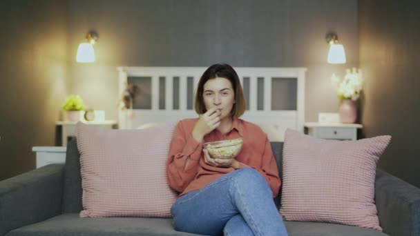 Happy young woman sitting on the sofa, eating popcorn, watching TV and laughing - 映像、動画