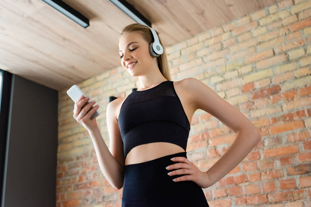 low angle view of happy sportswoman in wireless headphones using smartphone and standing with hand in pocket  - Fotografie, Obrázek