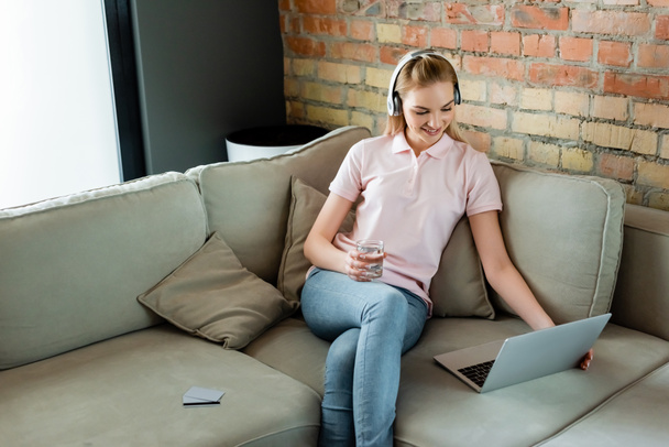 beautiful girl in wireless headphones looking at laptop and holding glass of water near credit cards on sofa - Foto, immagini