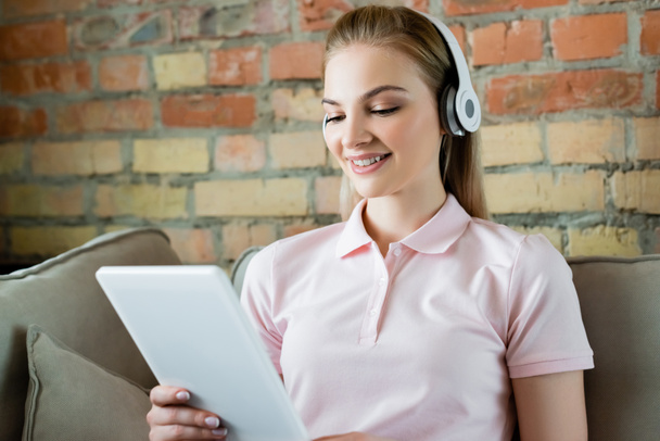 mujer sonriente en auriculares inalámbricos utilizando tableta digital en la sala de estar
  - Foto, Imagen