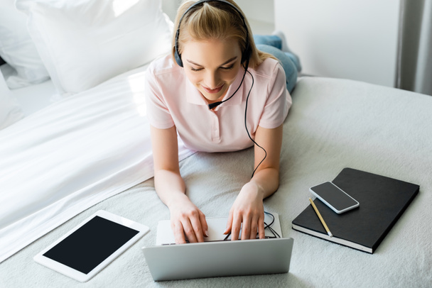 happy freelancer in headset using laptop near gadgets with blank screen on bed  - Fotografie, Obrázek