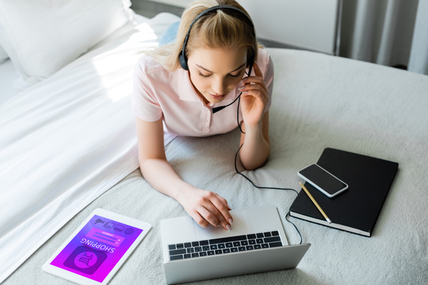 jonge freelancer in headset met laptop in de buurt van digitale tablet met shopping app en smartphone met leeg scherm op bed  - Foto, afbeelding