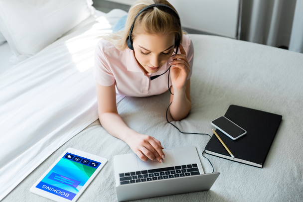 joven freelancer en auriculares con portátil cerca de tableta digital con aplicación de reserva y smartphone con pantalla en blanco en la cama
  - Foto, Imagen