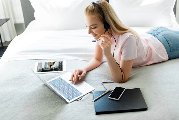 wesoła dziewczyna w zestawie słuchawkowym za pomocą laptopa w pobliżu tabletu cyfrowego z rezerwacją aplikacji i smartfona z pustym ekranem na łóżku  - Zdjęcie, obraz