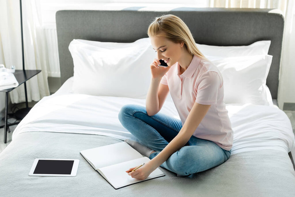 glückliche Frau schaut auf Notizbuch und spricht auf Smartphone neben digitalem Tablet mit leerem Bildschirm im Schlafzimmer  - Foto, Bild