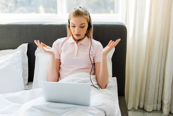 confused freelancer in headset showing shrug gesture near laptop in bedroom  - Fotografie, Obrázek