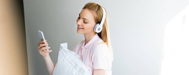 panoramiczne ujęcie uśmiechniętej kobiety w słuchawkach bezprzewodowych za pomocą smartfona w domu - Zdjęcie, obraz