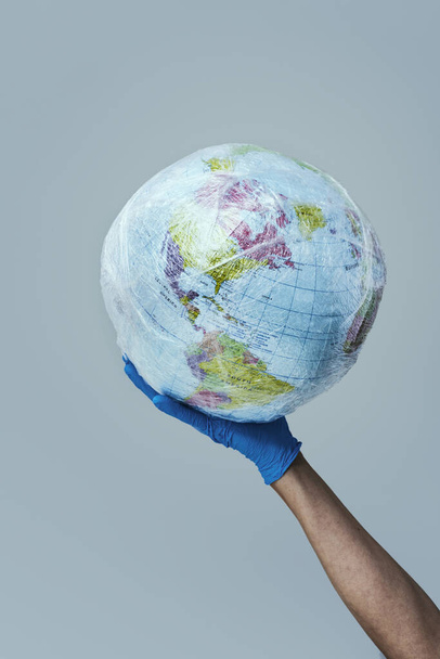 doctor man wearing blue surgical gloves holding a world globe wrapped in plastic, depicting the plastic contamination, or the protection against the epidemic infectious diseases or the air pollution - Photo, image