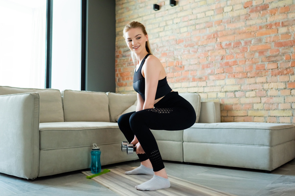 gelukkige sportvrouw doet squat oefening met halters in de buurt van sport fles in de woonkamer  - Foto, afbeelding