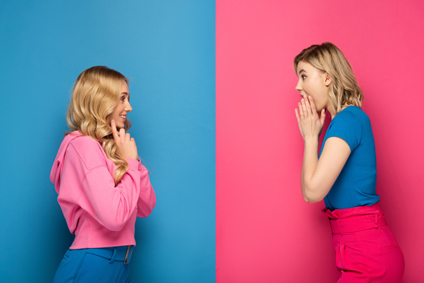 Seitenansicht schockierter und lächelnder blonder Schwestern, die einander auf rosa und blauem Hintergrund anschauen - Foto, Bild