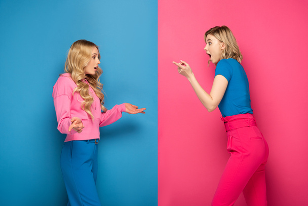 Side view of excited blonde sisters showing shrug gesture and pointing with finger on pink and blue background - Fotó, kép