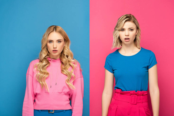 Attraktives blondes Mädchen, das in die Kamera schaut, in der Nähe einer schockierten Schwester auf rosa und blauem Hintergrund - Foto, Bild