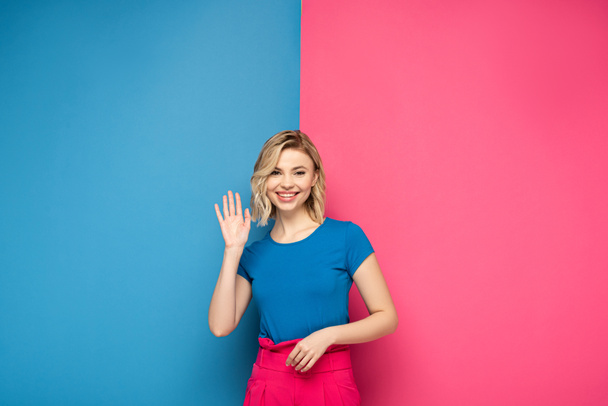 Smiling blonde woman waving hand at camera on pink and blue background - Fotó, kép