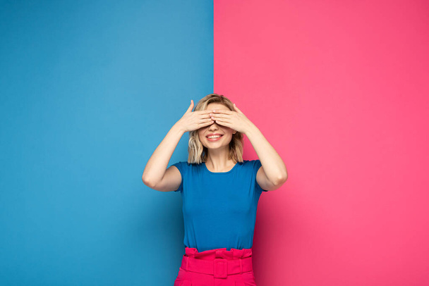 Smiling blonde girl covering eyes on pink and blue background - Foto, immagini