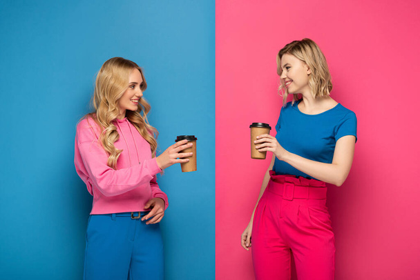 Positive blonde girls with paper cups looking at each other on pink and blue background - Фото, зображення