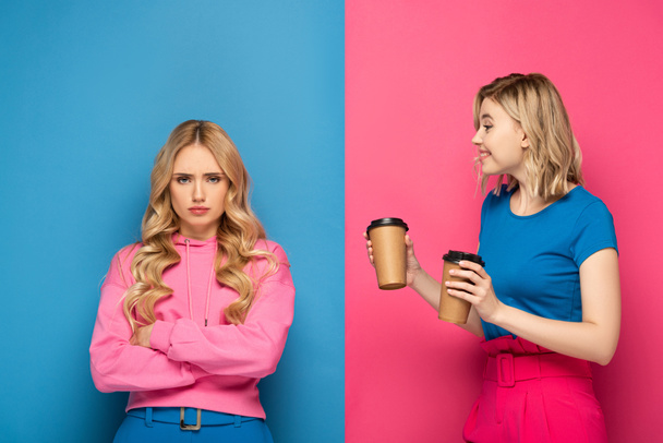 Smiling blonde girl with paper cups looking at offended sister on pink and blue background - Фото, изображение