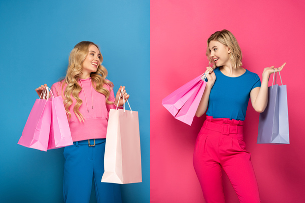 Smiling blonde girls with shopping bags looking at each other on pink and blue background - Fotoğraf, Görsel