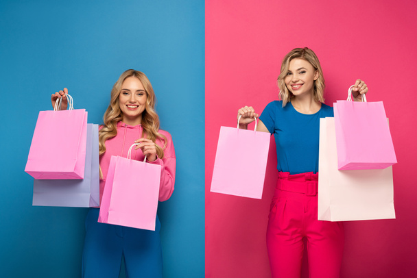 Mooie blonde zussen houden boodschappentassen en glimlachen op camera op roze en blauwe achtergrond - Foto, afbeelding