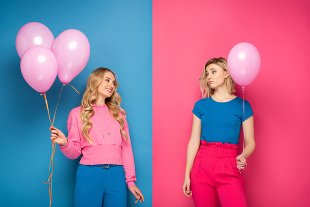 glimlachend meisje kijken naar boos blond zus met ballon op blauw en roze achtergrond - Foto, afbeelding
