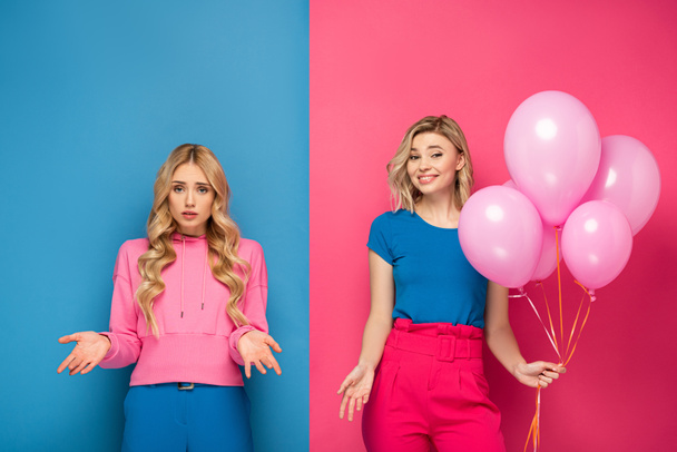 Smiling blonde girl holding balloons near offended sister on blue and pink background - 写真・画像