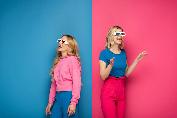 Smiling blonde sisters in 3d glasses looking away on blue and pink background - Fotó, kép