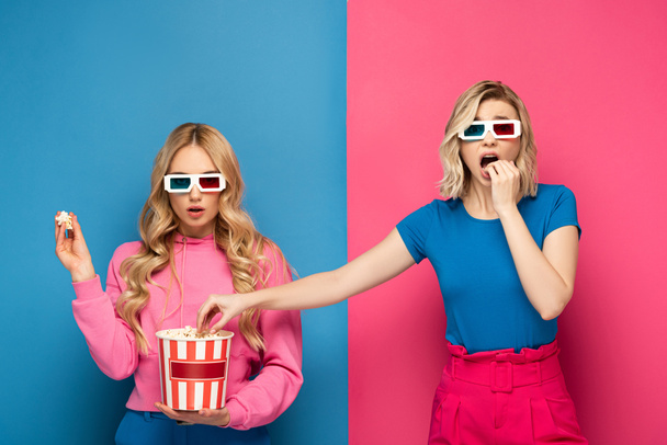 Attractive blonde girls in 3d glasses eating popcorn on blue and pink background - Fotografie, Obrázek